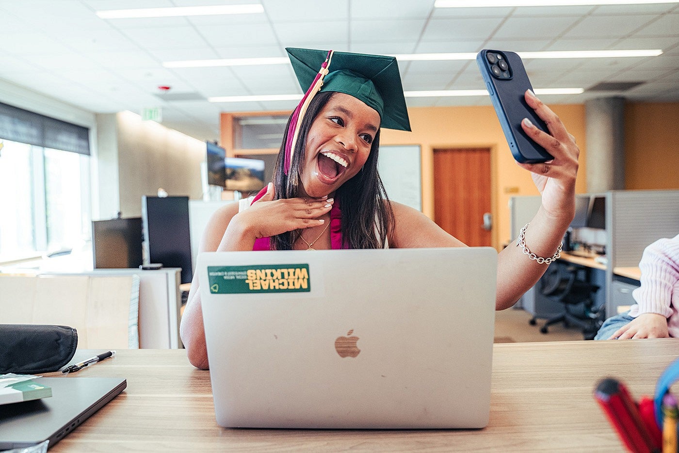 Student in a graduation cap in front of a laptop, smiling and taking a selfie.