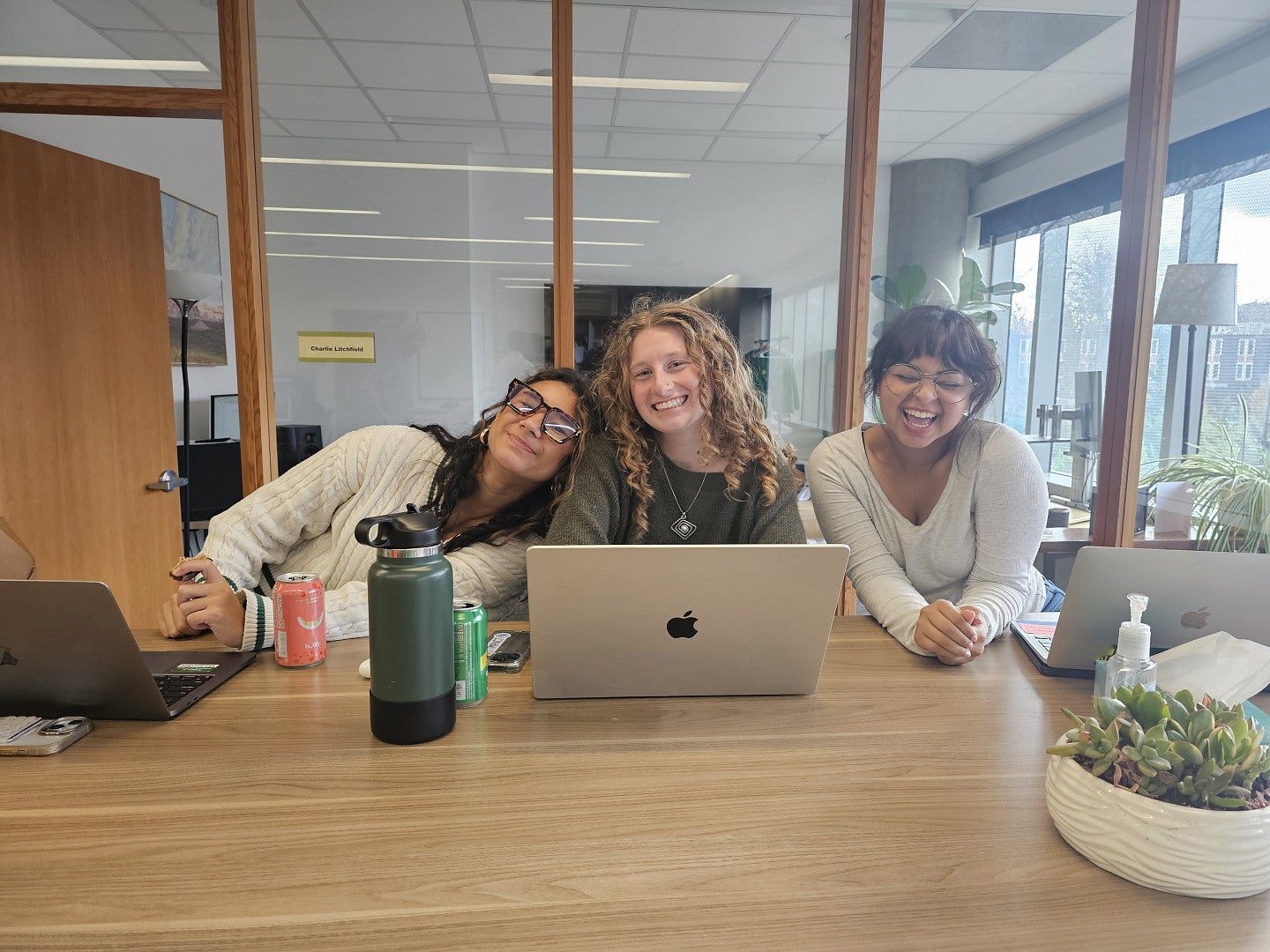 Three students sitting at a table smiling at the camera. There is a laptop and other miscellaneous items on the table.